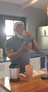 Dikey video: Kıdemli Afro-Amerikalı adam mutfakta smoothie yudumluyor telefon kaldırıyor ve selfie çekiyor. Sağlık, sağlık, yaşam tarzı, modern, samimi, rahatlama, dikey video