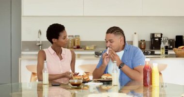 Çeşitli sosları ısırmak için hamburgeri kaldıran kadın mutfaktaki cam masada gülüyordu. Çift, yemek, mutfak, sosyal, günlük, canlı, yaşam tarzı