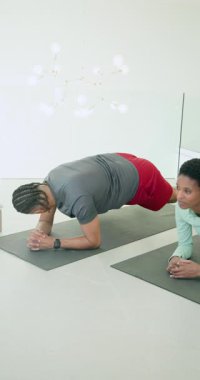 Dikey video: Kadın koç evdeki minderlerin üzerinde kobra esneme hareketlerine erkekleri tahta sürücüye yönlendiriyor. Fitness, kuvvet, sağlık, eğitim, aktif giyim, spor salonu, dikey video