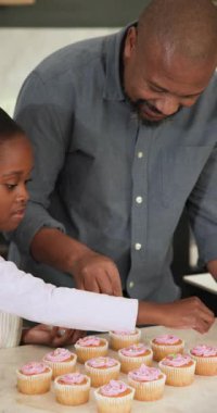 Dikey video: Dondurmadan sonra, baba ve kızı mutfakta eğlenmek için keklerin üzerine boncuk serpiyorlar. Aile, pişirme, kutlama, işçilik, samimiyet, bağ, dikey video