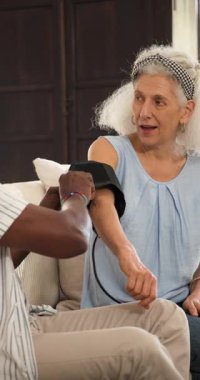 Kıdemli Afro-Amerikalı adam kelepçelerini takıyor, evde olgun beyaz kadın için kan basıncı okuyor. Sağlık, bakım, arkadaşlık, ev sağlığı, yaşlı bakımı, tıbbi teknoloji, dikey video