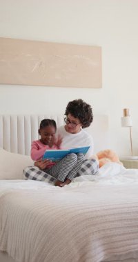 Dikey video: Afro-Amerikan anne kitap okuyor kızı yatakta işaret ediyor ve alkışlıyor. Aile, bağ, eğitim, yetiştirme, rahat, minimalist, dikey video