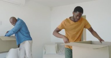 Afro-Amerikalı baba kutuyu açarken oğlu da oturma odasında flapları ve tişörtleri topluyor. Aile, yerli, gündelik, içsel, organizasyon, bağlanma, yaşam tarzı