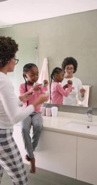 Dikey video: Afrikalı Amerikalı anne fırça seçiyor kızına banyo lavabosunda fırçalamayı öğretiyor. Aile, bakım, hijyen, çocukluk, eğitim, yaşam tarzı, dikey video