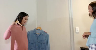 Yatak odasındaki ahşap askılarda tozlu pembe kazak ve kot gömlek tutan bayan arkadaşlar. Moda, tarz, gündelik, gençlik, yaşam tarzı, gardırop, modern.