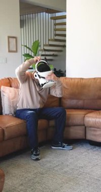 Dikey video: Adam VR kulaklığı alıyor, simülasyona başlıyor ve koltukla nesneleri yumrukluyor. Etkileyici, fütüristik, eğlence, modern, aktif, yaşam tarzı, dikey video