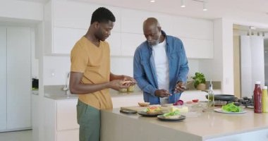 Afro-Amerikalı baba ve oğul sebze doğruyor. Mutfak adasına peynirli çörek ekliyorlar. Aşçılık, işbirliği, ev, beslenme, aile, sıcaklık, yaşam tarzı