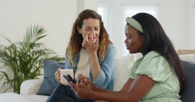 Kanepede oturan, akıllı telefon çalan, tepki veren ve kupayı yudumlayan çeşitli bayan arkadaşlar. Yoldaşlar, bağlantı, rahatlık, konfor, etkileşim, yaşam tarzı, iletişim