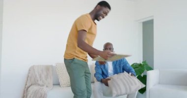 Afro-Amerikalı baba ve oğulun kutuları açması, kanepeye yastık yerleştirmesi. Aile, taşınma, ev dekorasyonu, çağdaş, yerli, bağlanma, yaşam tarzı