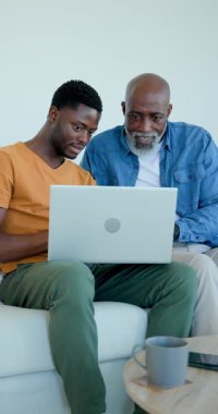 Dikey video: Afro-Amerikalı baba ve oğul kanepe üzerinde dizüstü bilgisayarı açıyor ve daktilo eden babaya yol gösteriyor. Aile, akıl hocalığı, teknoloji, öğrenme, bağlanma, gündelik, dikey video