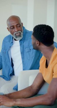 Dikey video: Afrikalı Amerikalı baba ve yetişkin oğul kanepede konuştuktan sonra rahatlığı paylaşıyorlar. Aile, danışmanlık, empati, iletişim, duygusal destek, bağlantı, dikey video