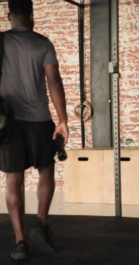 Dikey video: Spor salonuna giriliyor, Afrikalı Amerikalı bir adam spor çantasını bırakıyor ve ısınmak için esniyor. Fitness, antrenman, güç, kondisyon, kentsel, dikey video