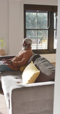 Dikey video: Afro-Amerikalı bir adam evde bilgisayarını yerleştiriyor iş e-postaları yazıyor ve dışarıya bakıyor. Üretkenlik, rahatlama, çalışma alanı, rahat, doğal ışık, modern, dikey video