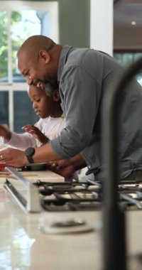 Dikey video: Siyah baba, kek tepsisini kaplarken kızının mutfakta karıştırmasına rehberlik ediyor. Aile, akıl hocalığı, bağlanma, aile içi, yaşam tarzı, samimiyet, dikey video