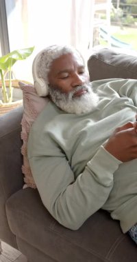 Dikey video: Yaşlı Afro-Amerikan adam, gevşemek için koltukta kulaklık takıyor. Rahatlama, rahatlık, rahatlık, modern, teknoloji, sükunet, dikey video