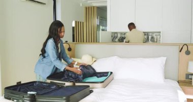 Afro-Amerikalı çift parlak yatak odasına giriyor ve seyahat için açık bavula giysilerini yerleştiriyor. Çift, paketleme, organizasyon, seyahat, yaşam tarzı, minimalist, modern