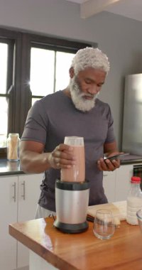 Dikey video: Kıdemli adam telefonda yemek tarifi okuyor, mutfakta blender 'da smoothie hazırlıyor. Sağlıklı, beslenme, modern, mutfak, mutfak, yaşam tarzı, dikey video