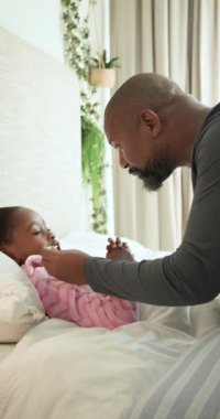 Dikey video: Afro-Amerikalı bir adam yatakta termometreyle kızın ateşini ölçüyor, kopyalama uzayı. Bakıcı, aile, sağlık, ev, beslenme, konfor, dikey video