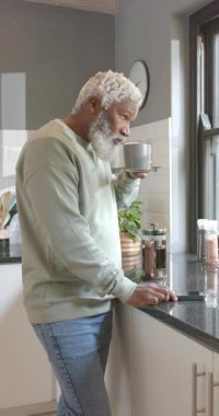 Dikey video: Yaşlı Afrikalı Amerikalı adam telefona bakıyor ve mutfak penceresinde kahve yudumluyor. Düşünce, rahatlama, sade, sıcak, gündelik, huzurlu, dikey video