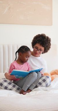 Çeşitli anne ve kız çocukları yatakta mavi kitap okuyor ve kızları resimlerin izini sürmeye teşvik ediyor. Aile, bağ, eğitim, hikaye anlatma, konforlu, besleyici, dikey video