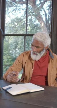 Dikey video: Kıdemli Afro-Amerikalı adam tableti tıklatıyor sonra da ev ofisinde deftere yazıyor. Profesyonel, çalışma alanı, modern, teknoloji, konsantrasyon, üretkenlik, dikey video