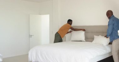 Afro-Amerikalı baba ve oğul yatak odasının kapısını açıyor, yastıkları kabartıyorlar ve rahat ettirmek için hizalıyorlar. Aile, takım çalışması, aile içi, uyum, rahat, ev dekorasyonu, kaynaşma.