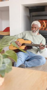 Dikey video: Kıdemli Afro-Amerikan adam tıngırdayan akorlar çalıyor, gitarın tonunu şömine başına ayarlıyor. Rahatlama, sanatçılık, akustik, konforlu, ambians, işçilik, dikey video