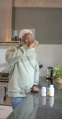 Dikey video: Cam kaldırma, kıdemli Afro-Amerikan erkek suyu yudumlama, mutfağa yansıma. Sağlık, farkındalık, huzur, tefekkür, zarafet, yaşam tarzı, dikey video