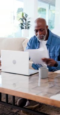 Dikey video: Yetişkin Afro-Amerikalı adam dosyaları okuyor, evde dizüstü bilgisayarda yazıyor. Yönetici, konsantrasyon, çalışma alanı, teknoloji, profesyonellik, üretkenlik, dikey video