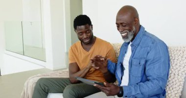 Afro-Amerikalı baba ve oğul tableti işaret ediyor, sürprizi buluyor, kanepede kahve fincanından yudumluyor. Aile, bağ, teknoloji, iletişim, rahatlama, sıcaklık, yaşam tarzı