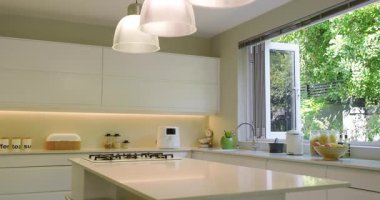 Pencereden süzülen güneş ışığı, mutfak adasında dönen kamera, meyve kasesi ve bahçe manzarası. Modern, minimalist, parlak, dingin, doğal, samimi, işlevsel