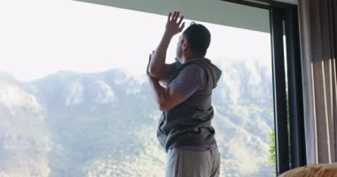 Asyalı adam, yoga minderini açıp esneyerek sürgülü cam kapıdaki sabah güneşine tepki veriyor. Sağlık, açık hava, spor, rahatlama, huzur, manzara, yaşam tarzı.