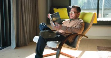 Asyalı olgun bir adam Sonsuz Günbatımlarını okuyor. Gözlükleri siliyor. Sandalyede rahatlamak için pencere pencere geziyor. Düşünce, rahatlık, sükunet, okuma, modern, iç mekan, yaşam tarzı