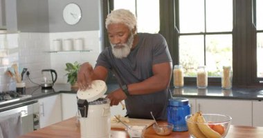 Kıdemli Afro-Amerikalı adam blender alıyor, smoothie yapıyor ve mutfak adasında muz dilimliyor. Sağlıklı, beslenme, yaşam tarzı, modern, ev yapımı, aşçılık, sağlık