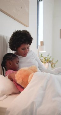 Anne ve kızı battaniyenin altında, Teddy kitap açıp sesli kitap okuyarak uyumaya yardımcı oluyor. Aile, yakınlık, yatma zamanı, beslenme, rahatlık, hikaye anlatma, dikey video