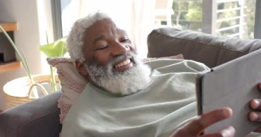 Kanepeye yaslanmış Afro-Amerikalı son sınıf öğrencisi tableti tıklatıyor ve evde kitap okumak için konuşuyor. Son sınıf, rahatlama, teknoloji, eğlence, iç mekan, yaşam tarzı, konfor.