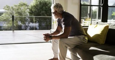 Yaşlıca Afrikalı Amerikalı bir adam oturma odasına giriyor, kanepeye oturuyor, dışarıya bakıyor, kameraya gülümsüyor. Düşünce, rahatlama, dinginlik, modern, sıcak, doğal, boş zaman