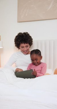 Tableti eğdikten sonra, çeşitli anne, anaokulu kızına yatakta kitap okuması için rehberlik ediyor. Bakıcı, etkileşim, şefkat, teknoloji, eğitim, konforlu, dikey video