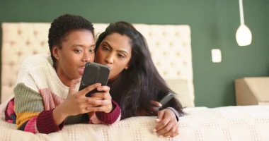 Siyah kadın akıllı telefonları kaydırıyor ve Hintli kadın pofuduk yatağı işaret ediyor ve gülüyor. Arkadaşlık, rahat, rahat, neşeli, hayat dolu, yaşam tarzı, birliktelik