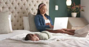 Kameralar dizüstü bilgisayarda çalışan bir kadının battaniyenin üzerinde uyuyan bebeğe bakarken görüntülerini çekiyor. Aileler, rahatlama, rahatlık, ev dekorasyonu, kadınlık, gündelik, çağdaş