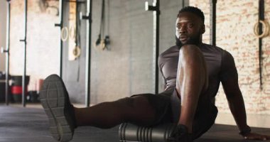 Afrikalı Amerikalı erkek spor salonunda sol baldırın altına köpük paten yerleştiriyor ve gerilim atmak için yuvarlanıyor. Fitness, hareketlilik, iyileşme, sağlık, güç, terapi, gerilme
