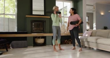 Yoga minderleri tutmak, son sınıf arkadaşlarının sohbet etmesi ve oturma odasında kameraya doğru yürümesi. Sağlık, dostluk, rahatlama, yaşam tarzı, modern, gündelik, arkadaşlık