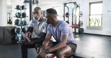 Afrikalı Amerikalı antrenör tableti tıklatıyor, müşterileri spor salonunda izlerken rutini açıklıyor. Spor, eğitim, sağlık, koçluk, motivasyon, teknoloji, güç
