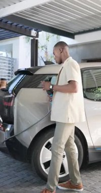 Afro-Amerikalı bir çift kahveyle eve geliyor, şarj etmek için EV şarj aletine bağlanıyorlar, bardaklarını paylaşıyorlar. Sürdürülebilirlik, yeşil enerji, elektrikli araç, modern yaşam tarzı, rahatlama, açık hava, dikey video