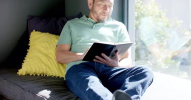 Olgun Asyalı adam kanepenin yanındaki kanepede yazı yazıyor, güneşli yapraklara bakmak için duruyor. Konsantrasyon, yansıma, sükunet, ilham, evcil, doğal, minimalist