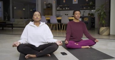 Hintli ve Afrikalı Amerikalı kadınlar akıllı telefonun yanında yoga minderleri üzerinde meditasyon yapıyorlar, gülümsemelerini paylaşıyorlar. Farkındalık, sağIık, sükunet, bağlantı, arkadaşlık, çağdaş, iç mekan