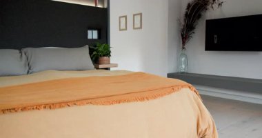 Sahne açılışı, turuncu atımlı ve gri yastıklı yatağın yanından geçen kamera ile başlıyor. Minimalist, çağdaş, iç mekan, dekor, yaşam tarzı, sakinleştirici, tarafsız