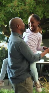 Dikey video: Kızını kaldıran adam bağlanmak için gölgeli bahçe avlusunda kıvrılıp sallanıyor. Şefkat, bağ, aile, açık hava, boş zaman, mutluluk, dikey video