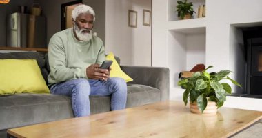 Kıdemli Afro-Amerikan adam, evde okuma uyarısı ve sandık başında, koltukta akıllı telefon kaydırıyor. Sağlık, sağlık, teknoloji, rahatlama, iç mekan, yaşam tarzı, konfor