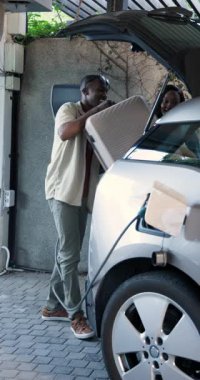 Afro-Amerikalı çift EV 'yi ev garajının kaputunda şarj ediyor, kabloları bağlıyor, ödüyor, çıkıyor. Sürdürülebilirlik, yenilik, modern, teknoloji, çevre dostu, yaşam tarzı, dikey video